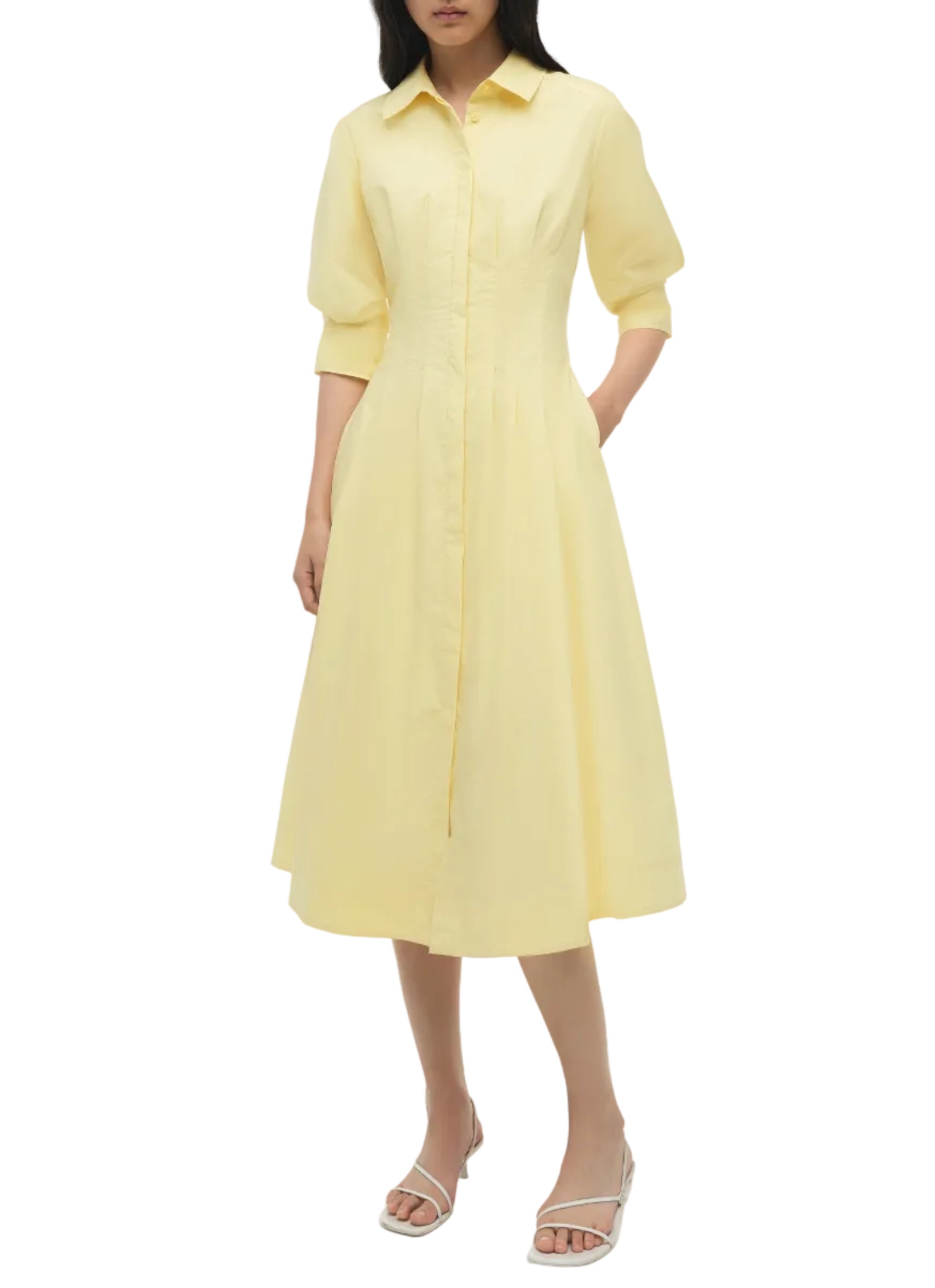 A woman wears the Simkhai Jazz Pintuck Midi Dress in light yellow cotton, featuring a collared neckline, A-line silhouette, button-down front, elbow-length sleeves, and pockets, paired with white heeled sandals.