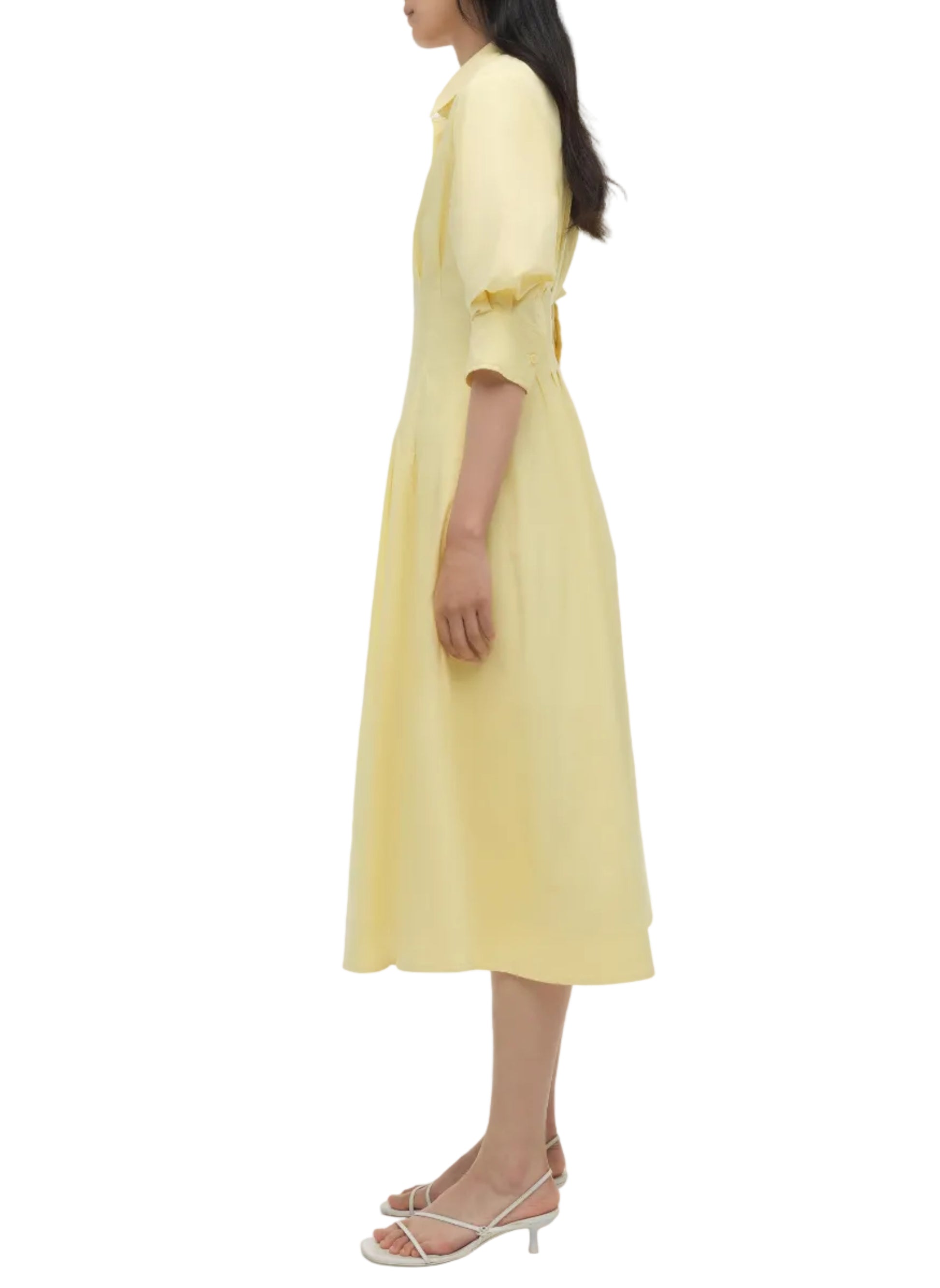 A woman in profile wears the Simkhai Jazz Pintuck Midi Dress, a light yellow cotton A-line style with elbow-length sleeves, paired with white strappy heeled sandals.