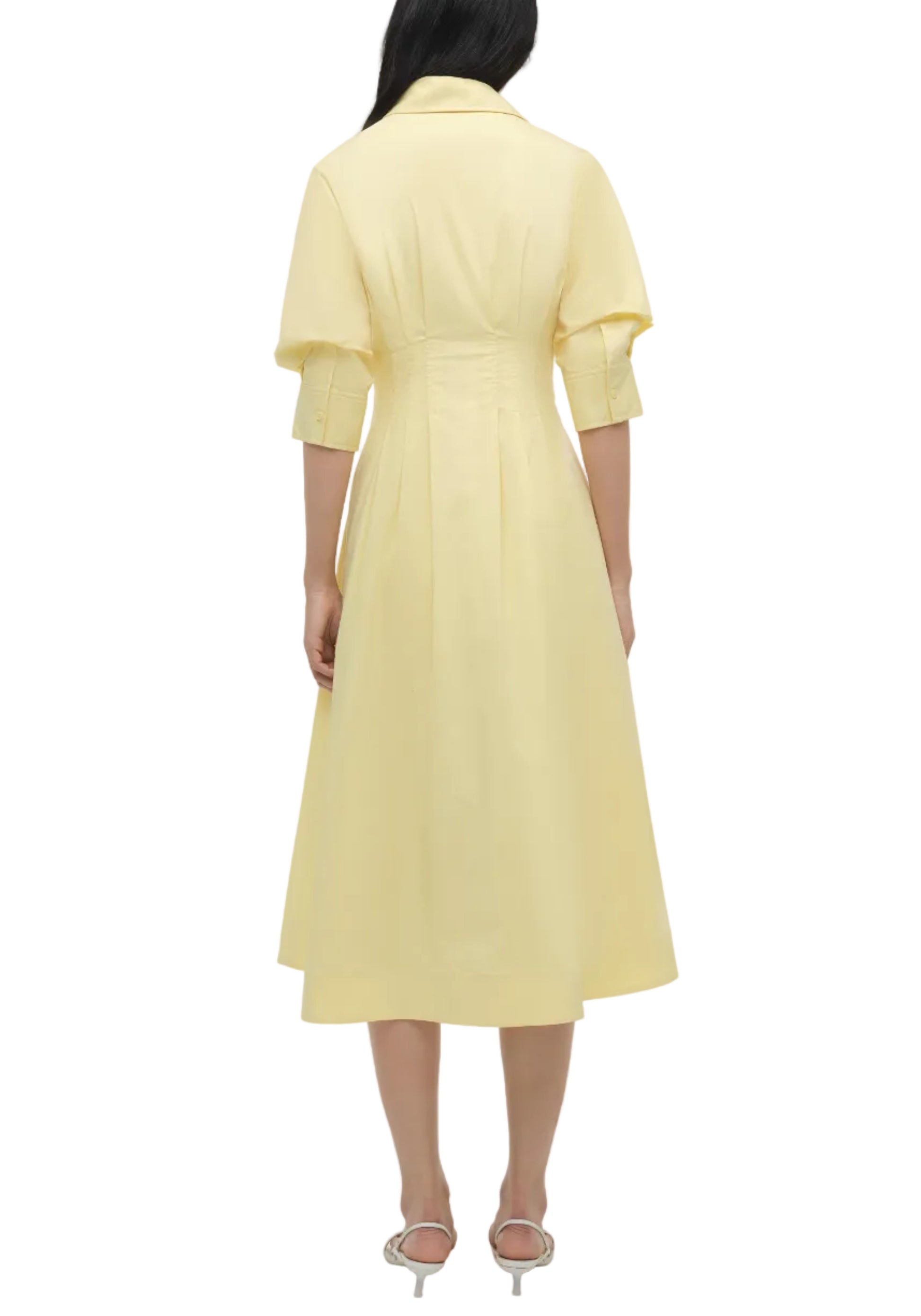 A woman is seen from behind wearing the Simkhai Jazz Pintuck Midi Dress in light yellow, featuring elbow-length sleeves, a collar, and an A-line silhouette. She pairs it with white heeled sandals.