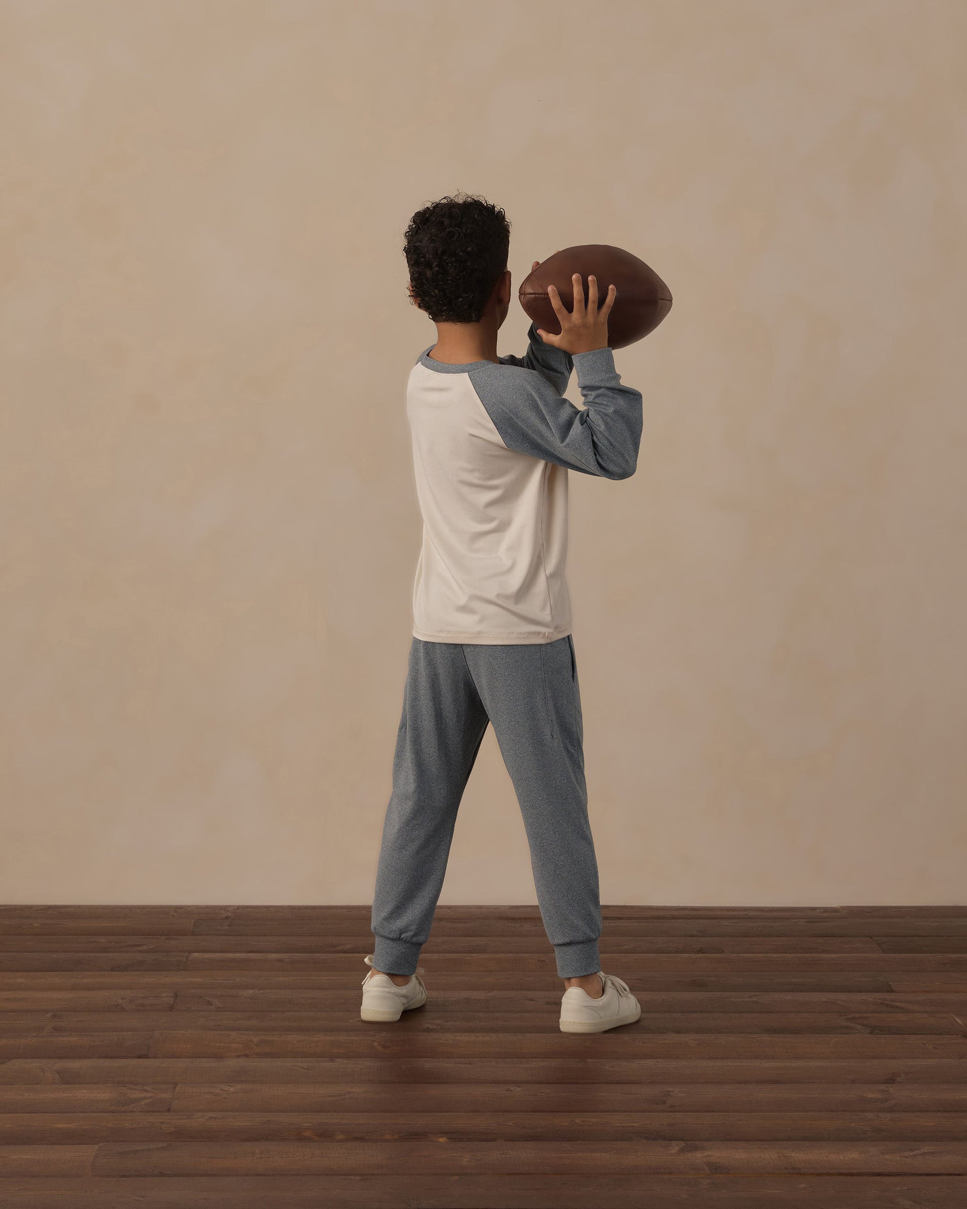 A child, dressed in the Rylee & Cru Boys’ Long Sleeve Raglan Tee pajamas, stands on a wooden floor holding a football and facing away from the camera against a beige wall, enjoying the breathable comfort of soft fabrics.