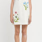 A woman models the Stella McCartney Floral Embroidered Mini Shift Dress, a sleeveless white dress adorned with vibrant floral embroidery, against a plain white background.