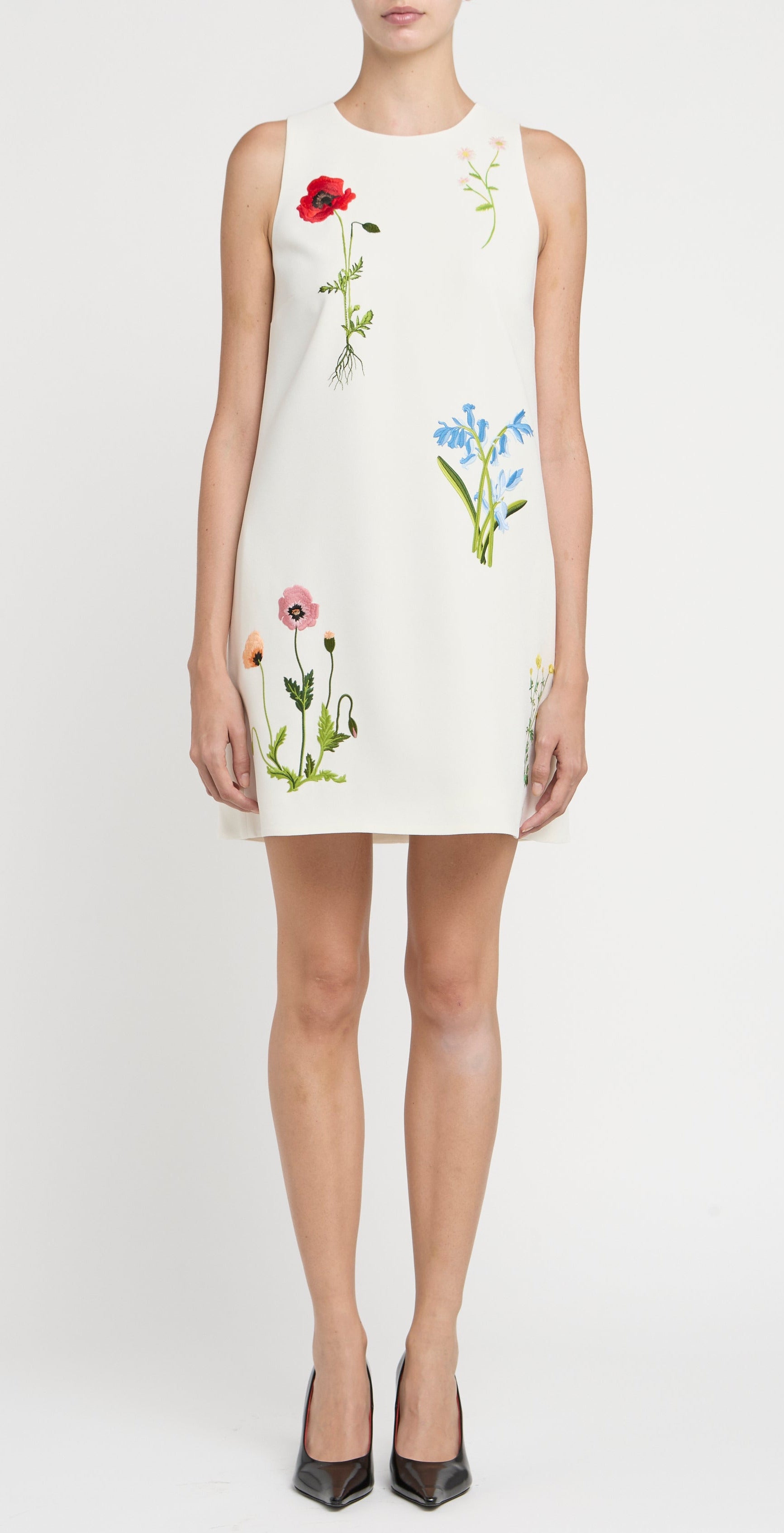 A woman models the Stella McCartney Floral Embroidered Mini Shift Dress, a sleeveless white dress adorned with vibrant floral embroidery, against a plain white background.