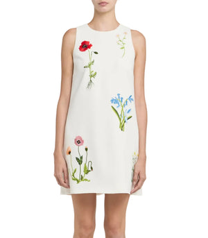 A woman wears the Stella McCartney Floral Embroidered Mini Shift Dress, a sleeveless white dress featuring colorful botanical flower embroidery, while standing against a plain background.
