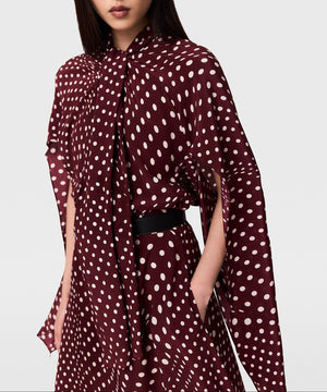 A person wears the Stella McCartney Tie-Neck Polka Dot Silk Dress—a maroon dress adorned with white polka dots, featuring a tie-neck detail and loose, flowing sleeves. The midi silhouette is cinched at the waist with a black belt, creating an effortlessly stylish look.