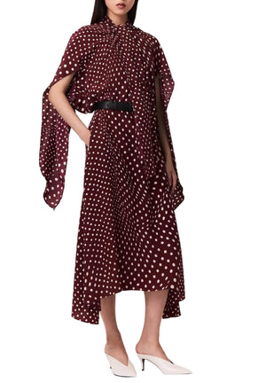 A woman models the Stella McCartney Tie-Neck Polka Dot Silk Dress, a maroon, ankle-length piece with a classic midi silhouette and tie-neck detail, styled with a black belt, white heels, and a draped cape accent, set against a white background.