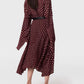 A person wears the Stella McCartney Tie-Neck Polka Dot Silk Dress in a classic maroon midi silhouette with long sleeves and a black belt, paired with white heeled mules, standing against a plain light background.