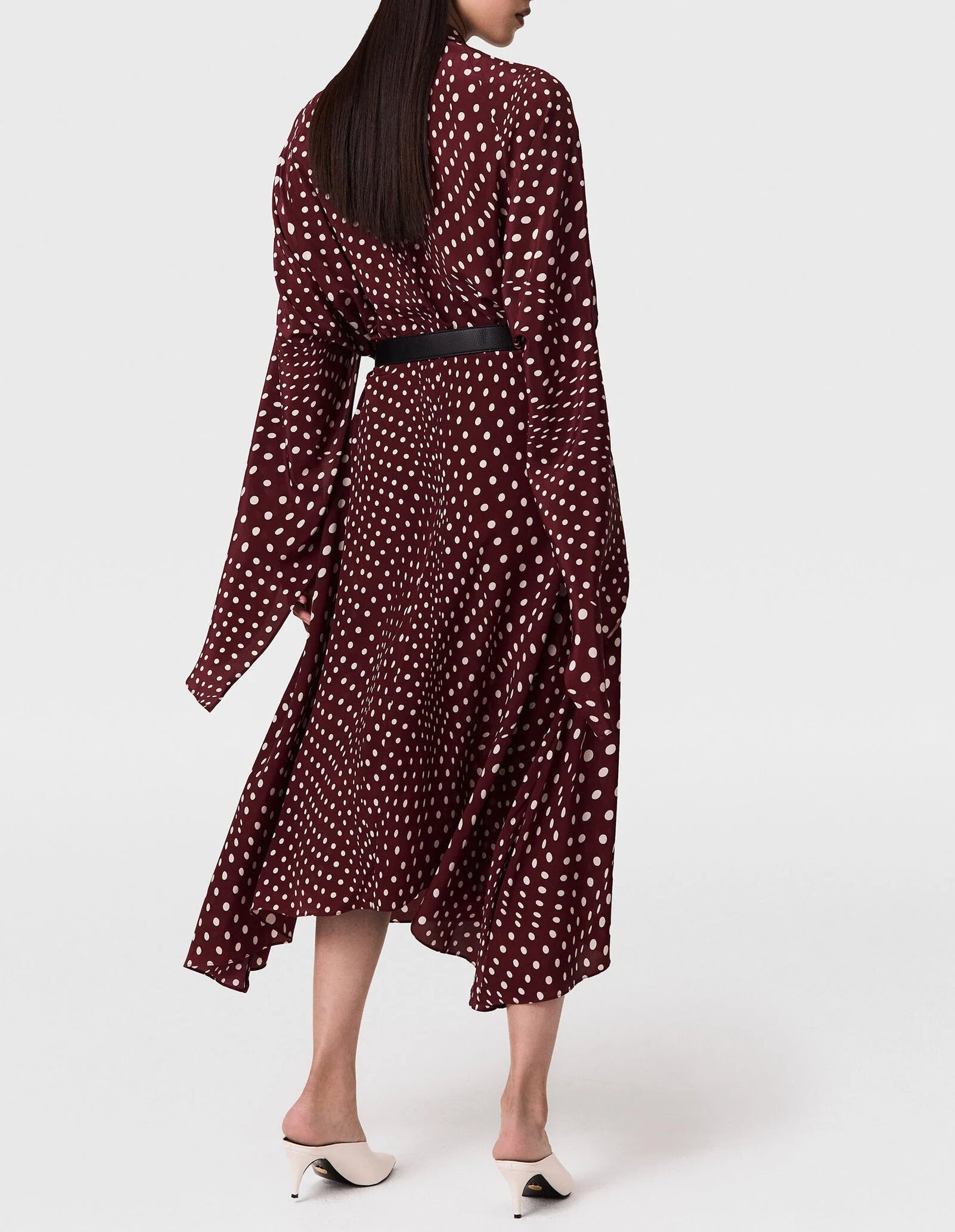 A person wears the Stella McCartney Tie-Neck Polka Dot Silk Dress in a classic maroon midi silhouette with long sleeves and a black belt, paired with white heeled mules, standing against a plain light background.