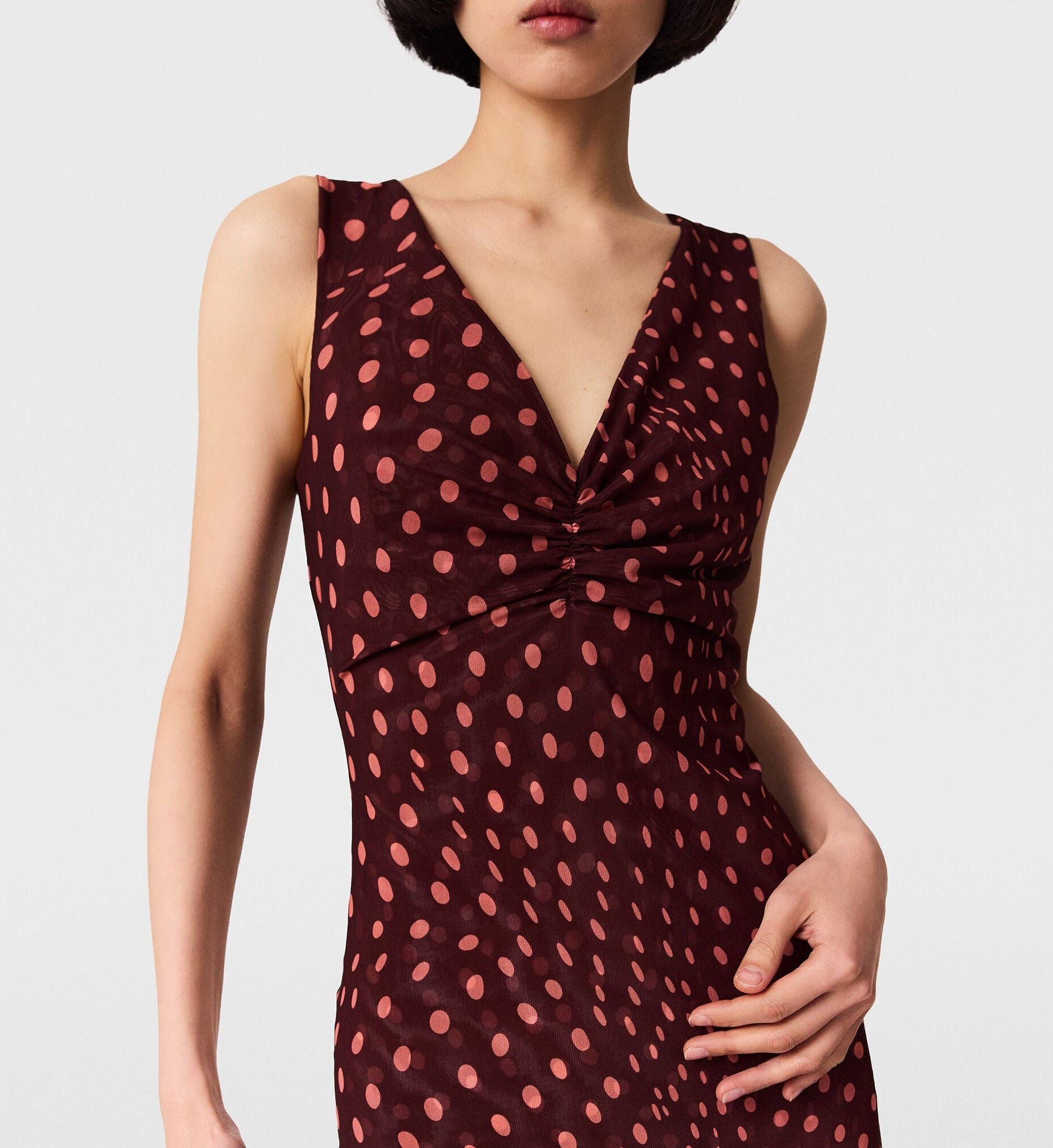 A woman wearing the Stella McCartney Polka Dot Mesh V-Neck Midi Dress, featuring a sleeveless design with maroon polka dots, pink accents, and a V-neckline, stands against a plain background.