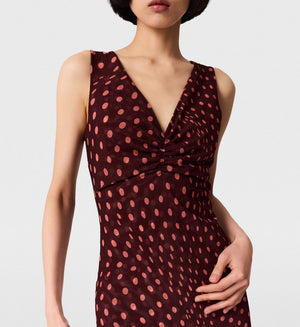 A woman wearing the Stella McCartney Polka Dot Mesh V-Neck Midi Dress, featuring a sleeveless design with maroon polka dots, pink accents, and a V-neckline, stands against a plain background.