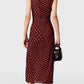A woman with a short bob haircut is seen from behind against a plain background, wearing the Stella McCartney Polka Dot Mesh V-Neck Midi Dress in maroon. The sleeveless dress features a flattering mesh silhouette adorned with polka dots. She pairs it with white heels and carries a small black handbag.