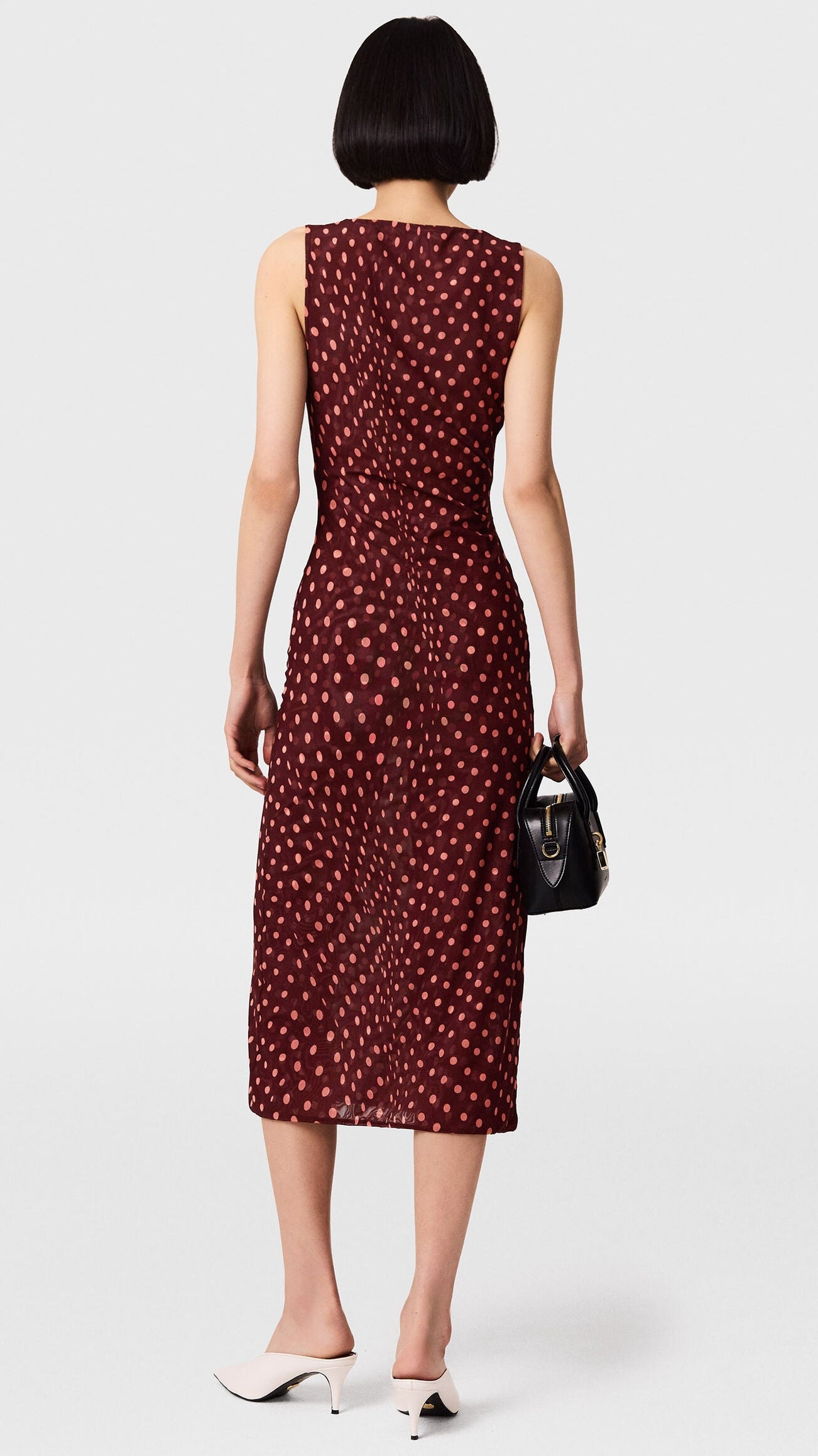A woman with a short bob haircut is seen from behind against a plain background, wearing the Stella McCartney Polka Dot Mesh V-Neck Midi Dress in maroon. The sleeveless dress features a flattering mesh silhouette adorned with polka dots. She pairs it with white heels and carries a small black handbag.