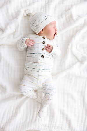 A newborn baby dressed in the Lou Lou & Company Baby Conrad Top & Bottoms, featuring a white design with dotted stripes and a matching hat, lies on a soft white blanket facing left. This cozy set is crafted from silky-soft fabric and includes built-in footies to keep your little one snug and comfortable.