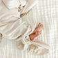 A newborn baby in the Baby Dakota Plush Knit Knotted Gown by Lou Lou & Company lies on a white quilted blanket, with a pacifier and hand near their face and feet visible—ideal for hassle-free diaper changes.