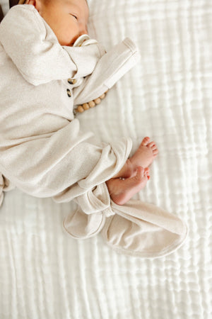 A newborn baby in the Baby Dakota Plush Knit Knotted Gown by Lou Lou & Company lies on a white quilted blanket, with a pacifier and hand near their face and feet visible—ideal for hassle-free diaper changes.