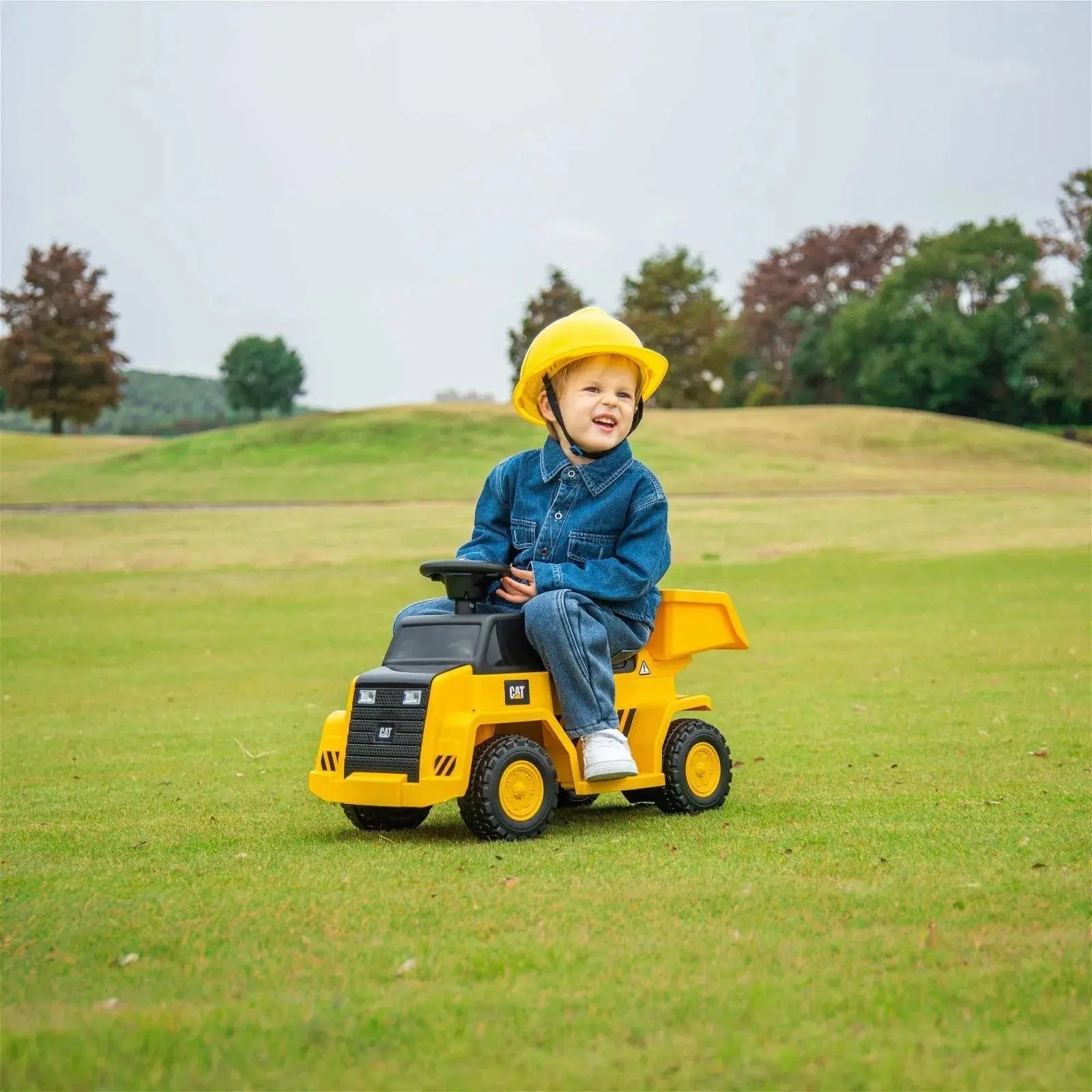 A young child wearing a yellow helmet and denim outfit sits on a Freddo 6V CAT Dump Truck Ride-On in a grassy field with trees in the background.
