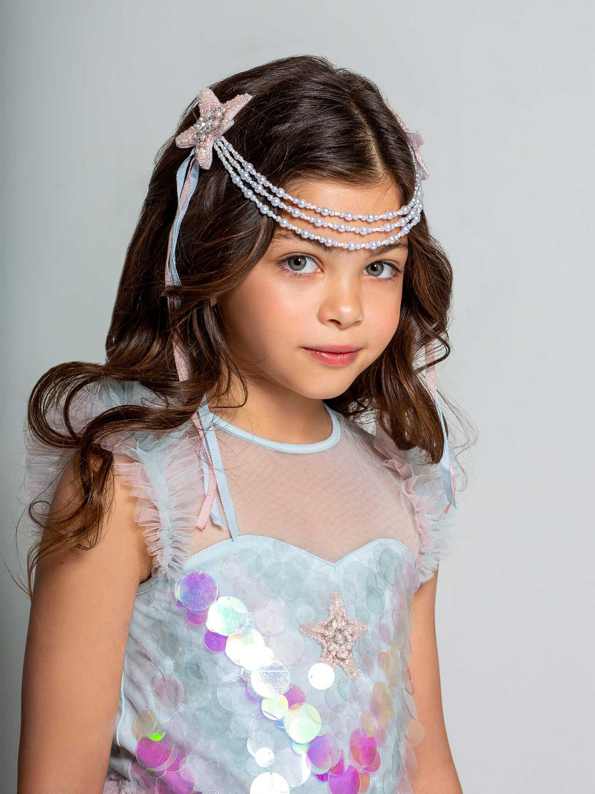 A young girl with long brown hair wears the Tutu du Monde Girls’ Mermaid Magic Tutu Dress, featuring sparkling sequins and a beaded headpiece, as she gazes at the camera against a simple light background, capturing the enchanting essence of a mermaid costume.