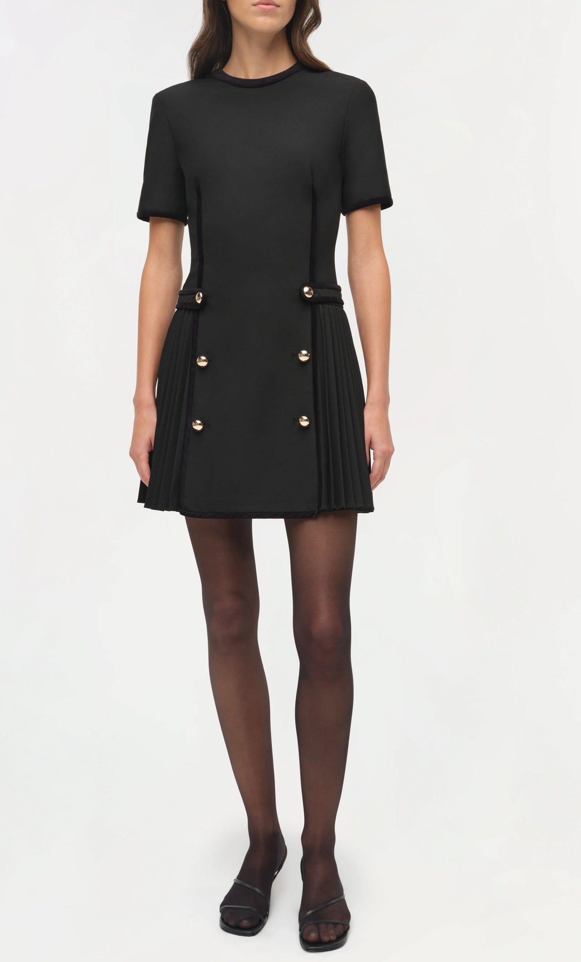 A woman wears the Simkhai Doria Mini Dress with gilded buttons, black sheer tights, and black open-toe heels, standing against a plain white background.