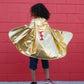 A child wearing sneakers and the Great Pretenders Gold & Red Reversible Wonder Cape, featuring a shiny gold side with a red lightning bolt, stands in front of a red brick wall. This reversible cape is proudly made in Canada.