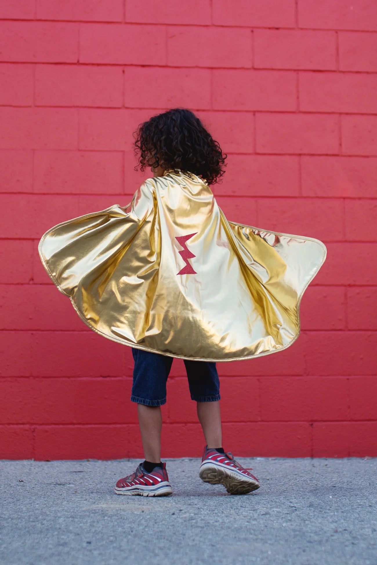 A child wearing sneakers and the Great Pretenders Gold & Red Reversible Wonder Cape, featuring a shiny gold side with a red lightning bolt, stands in front of a red brick wall. This reversible cape is proudly made in Canada.