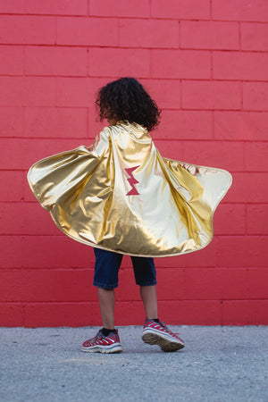 A child wearing sneakers and the Great Pretenders Gold & Red Reversible Wonder Cape, featuring a shiny gold side with a red lightning bolt, stands in front of a red brick wall. This reversible cape is proudly made in Canada.