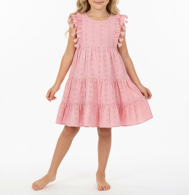 A young girl stands barefoot against a white background, wearing the Mer Girls’ Sophie Tassel Dress—a sleeveless, knee-length dress crafted from lightweight cotton with ruffle details and a tiered skirt.