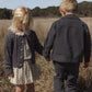 Two young children holding hands stand in a grassy field. The girl wears a dress and cardigan, while the boy sports the Rylee & Cru Baby Boys’ and Boys’ Chore Coat, featuring chest patch pockets—a classic layering piece. Their faces are partially turned away.