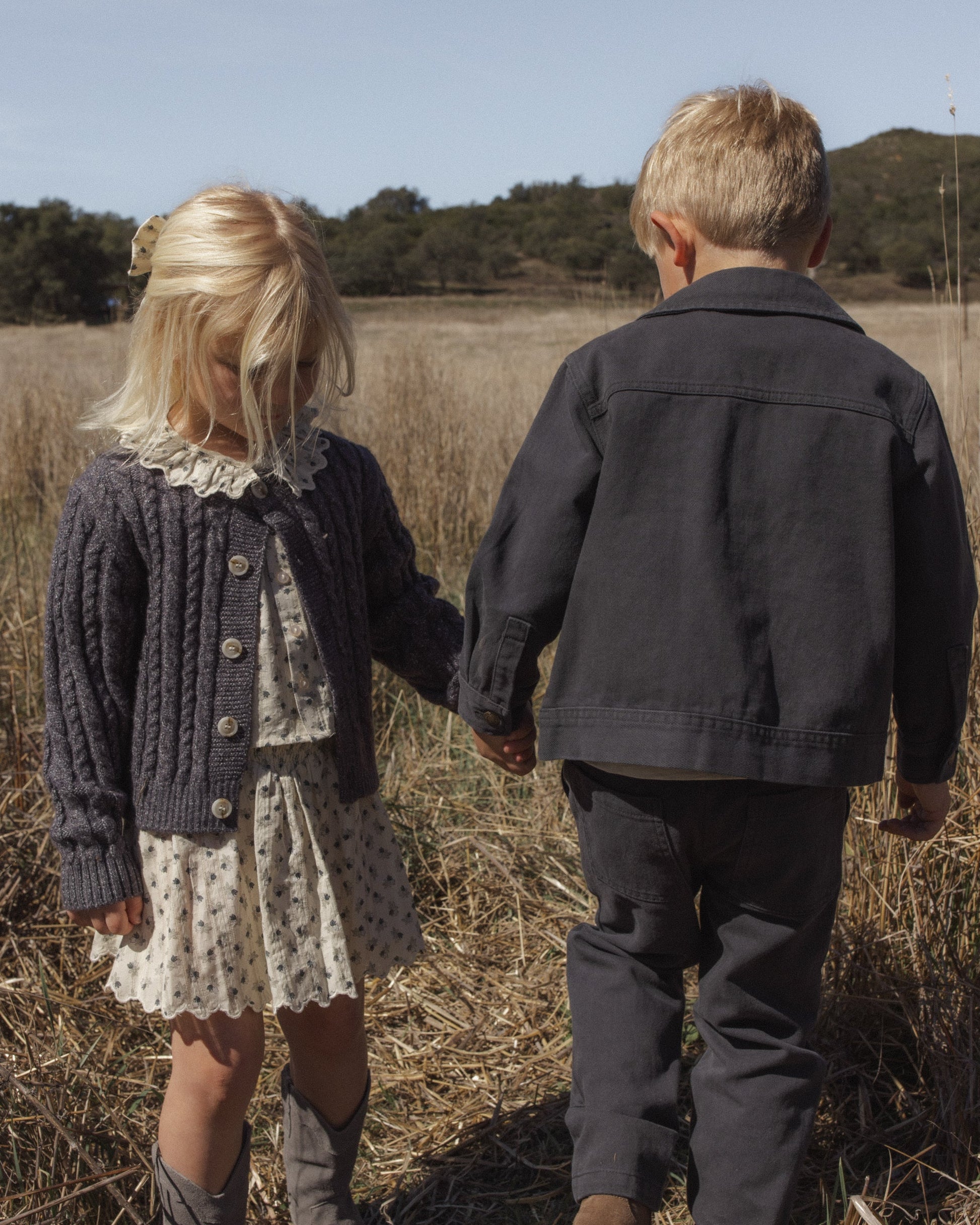Two young children holding hands stand in a grassy field. The girl wears a dress and cardigan, while the boy sports the Rylee & Cru Baby Boys’ and Boys’ Chore Coat, featuring chest patch pockets—a classic layering piece. Their faces are partially turned away.