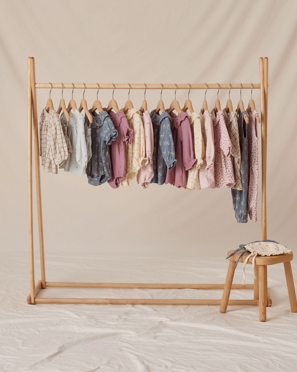 A wooden rack displays Quincy Mae Baby Girls' Collared Rompers with ruffle details and puff sleeves in neutral and pastel shades on hangers, accompanied by a small wooden stool and a cushion.