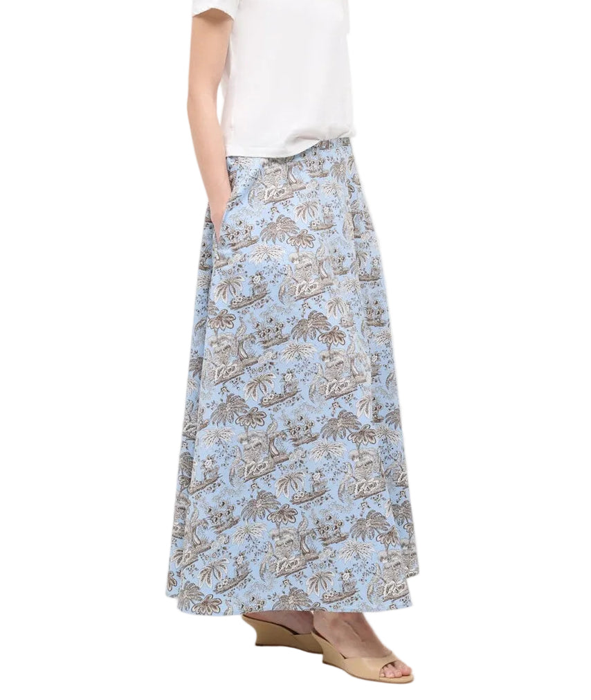A person wearing a white short-sleeve top, the Staud Axel Skirt with blue and beige floral patterns, and beige wedge sandals stands against a white background.