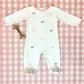 A Baby Girls' Rock Me Romper by The Beaufort Bonnet Company with delicate pink and purple bow embroidery rests on a pink checkered blanket beside a silver rattle.