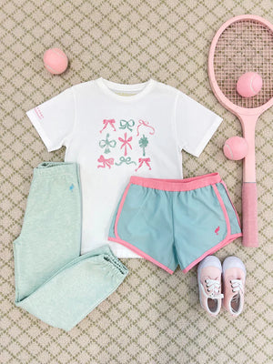 A white tee with bows, mint green joggers, Girls' Prepletic™ Shimmy Shake Shorts, pink and white sneakers, a pink tennis racket, and three pink tennis balls from The Beaufort Bonnet Company on a patterned carpet.