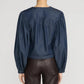 A person is shown from the back against a plain light background, wearing Frame’s The Off Duty Long Sleeve Popover—a dark blue lightweight denim blouse with a split neckline—paired with dark brown pants.