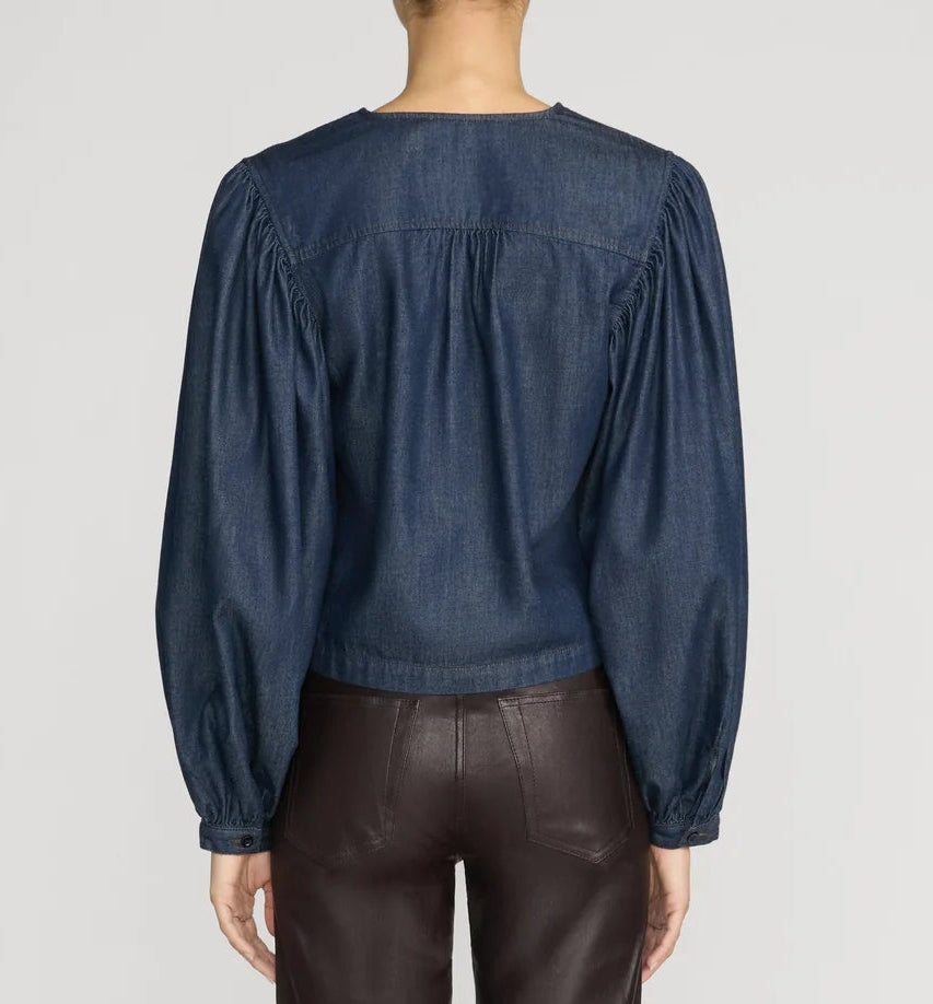 A person is shown from the back against a plain light background, wearing Frame’s The Off Duty Long Sleeve Popover—a dark blue lightweight denim blouse with a split neckline—paired with dark brown pants.
