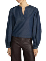 A woman is wearing The Off Duty Long Sleeve Popover by Frame, a dark blue denim blouse with a split V-neckline and gathered sleeves, paired with high-waisted dark brown leather pants.