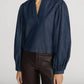 A person wearing The Off Duty Long Sleeve Popover by Frame—a long-sleeved, dark blue lightweight denim blouse with a split neckline and gathered sleeves—paired with dark brown leather pants, stands against a plain background.