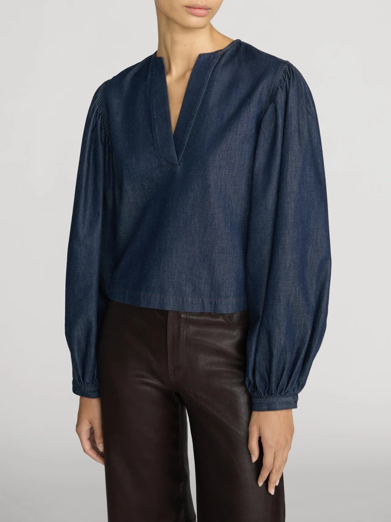 A person wearing The Off Duty Long Sleeve Popover by Frame—a long-sleeved, dark blue lightweight denim blouse with a split neckline and gathered sleeves—paired with dark brown leather pants, stands against a plain background.