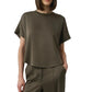 A person wearing the Simkhai Addy Satin T-Shirt, an olive green short-sleeve top with a classic round neckline, paired with matching high-waisted pants, stands with one hand in their pocket against a plain background. The relaxed fit of the ensemble lends effortless style and comfort.