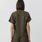 A person with straight, shoulder-length brown hair stands facing away, wearing the Simkhai Addy Satin T-Shirt in olive green—a short-sleeved top with a classic round neckline—paired with matching relaxed-fit pants, set against a plain light background.
