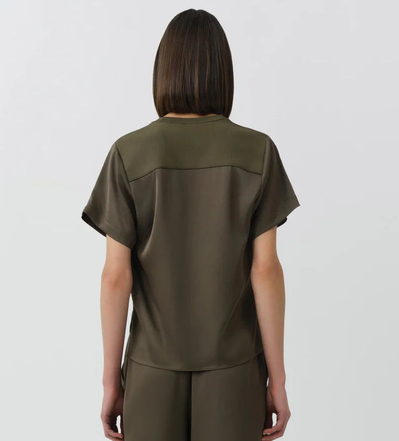 A person with straight, shoulder-length brown hair stands facing away, wearing the Simkhai Addy Satin T-Shirt in olive green—a short-sleeved top with a classic round neckline—paired with matching relaxed-fit pants, set against a plain light background.