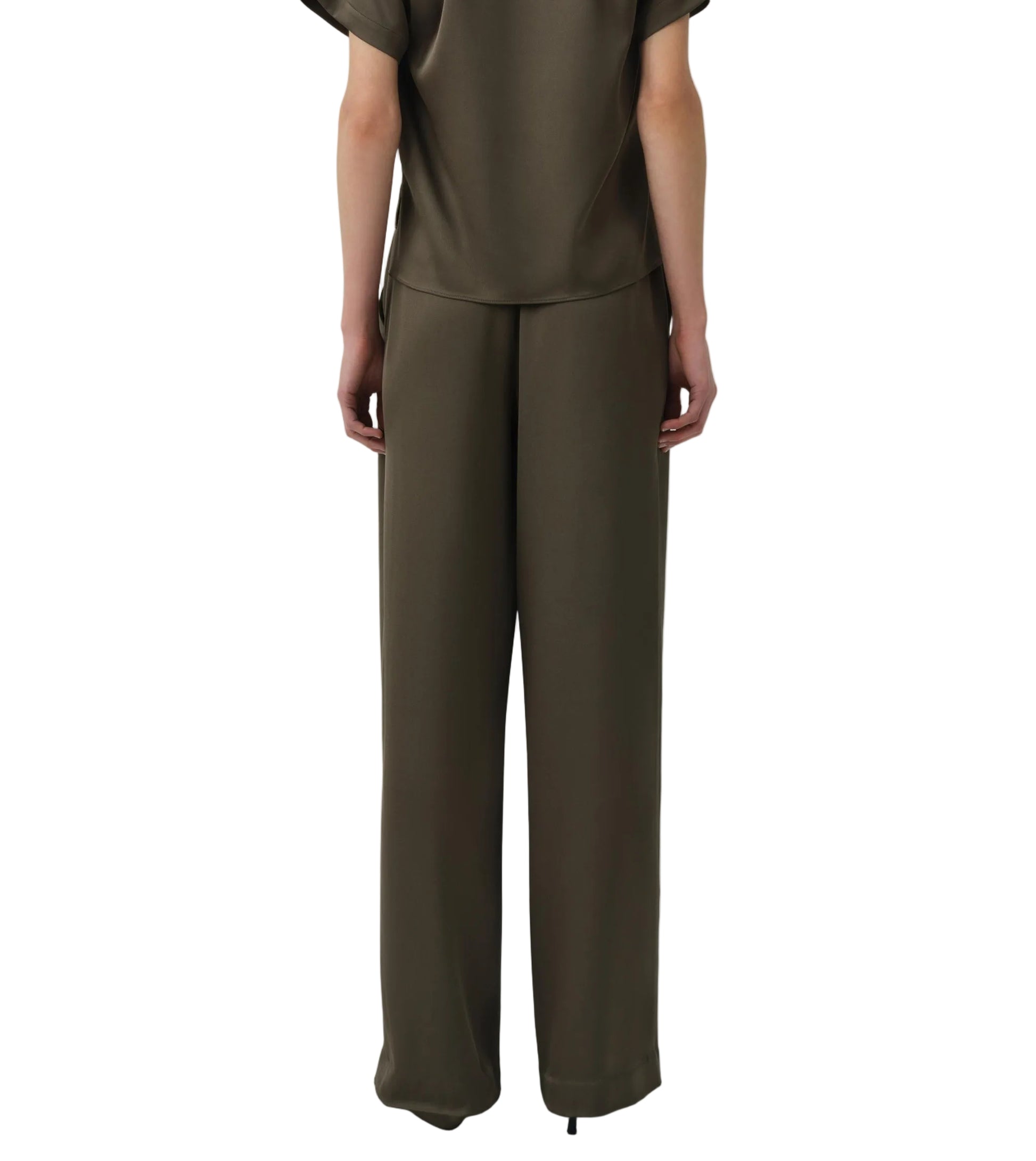 A person stands with their back to the camera, wearing Simkhai’s Kyra Wide Leg Pant in dark olive green satin with a tailored waistband and matching short-sleeve top for a relaxed silhouette.