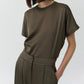 A woman wears the Simkhai Addy Satin T-Shirt in olive green—featuring a relaxed fit, short sleeves, and a classic round neckline—tucked into coordinating high-waisted trousers with her hands in the pockets, set against a plain light background.