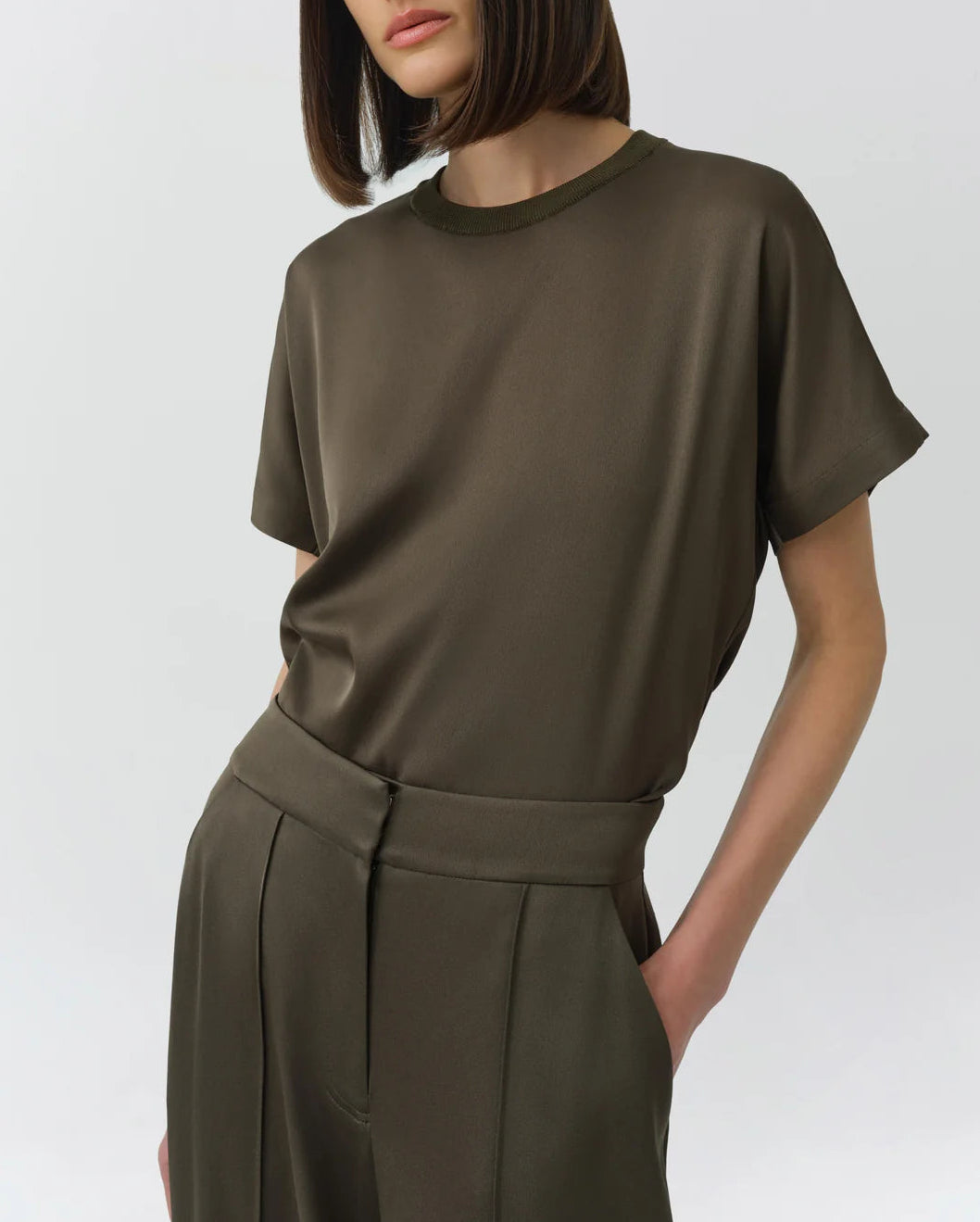 A woman wears the Simkhai Addy Satin T-Shirt in olive green—featuring a relaxed fit, short sleeves, and a classic round neckline—tucked into coordinating high-waisted trousers with her hands in the pockets, set against a plain light background.