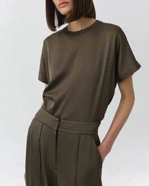 A woman wears the Simkhai Addy Satin T-Shirt in olive green—featuring a relaxed fit, short sleeves, and a classic round neckline—tucked into coordinating high-waisted trousers with her hands in the pockets, set against a plain light background.