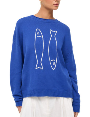 A woman stands against a plain background wearing the Staud Giada Sweater—a blue, lightweight summer sweater with two white pop sardines on the front—paired with a white skirt.