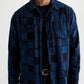 A man wearing the AG Jeans Louis Camp Shirt—a blue and black checker patchwork button-up cotton jacquard shirt—over a black t-shirt, paired with dark jeans and a visible black belt.