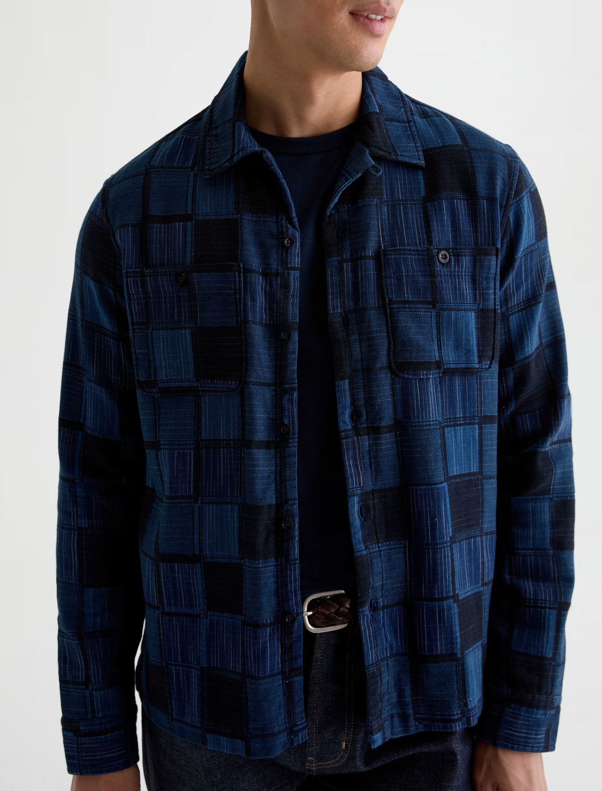 A man wearing the AG Jeans Louis Camp Shirt—a blue and black checker patchwork button-up cotton jacquard shirt—over a black t-shirt, paired with dark jeans and a visible black belt.