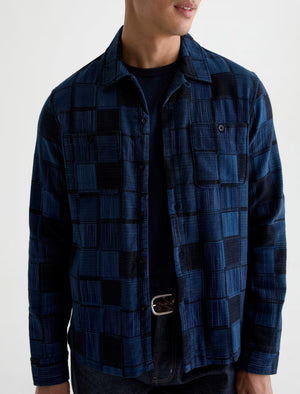 A man wearing the AG Jeans Louis Camp Shirt—a blue and black checker patchwork button-up cotton jacquard shirt—over a black t-shirt, paired with dark jeans and a visible black belt.