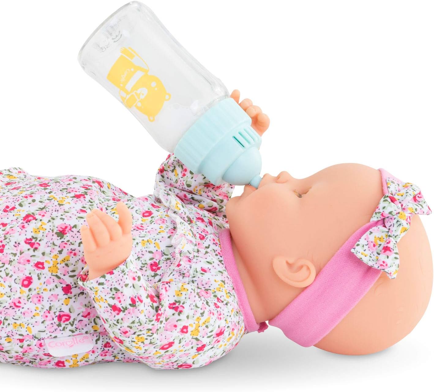 A baby doll wearing a floral outfit and pink headband is lying down, holding the Corolle Magic Milk Baby Doll Bottle for 14-17" to its mouth—the perfect feeding accessory.