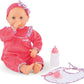 The Corolle Lila Chérie 17" baby doll wears a pink outfit and headband, sits with its thumb in its mouth next to a white bottle and matching pink bib.