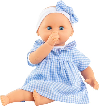 The Corolle Premier Calin - Milly Doll is a soft-bodied baby doll with blue eyes, a white headband with a bow, and a blue and white checkered dress, sitting with its thumb in its mouth.